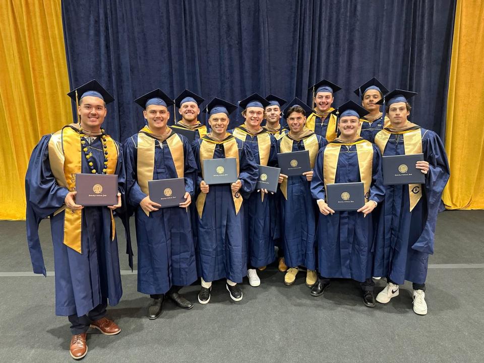 Twelve graduates in blue and gold caps and gowns stand together, holding diplomas and smiling in front of gold and navy curtains.