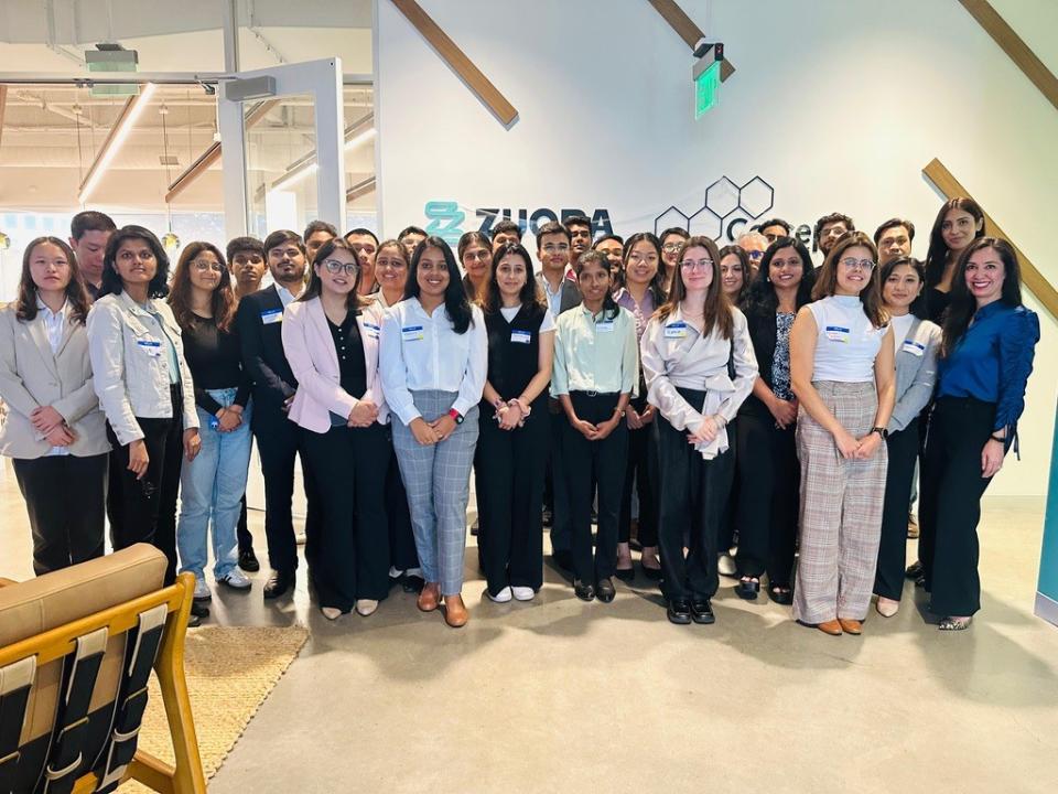 A large group of professionally dressed people pose for a group photo indoors in front of a wall with the Zuora logo.