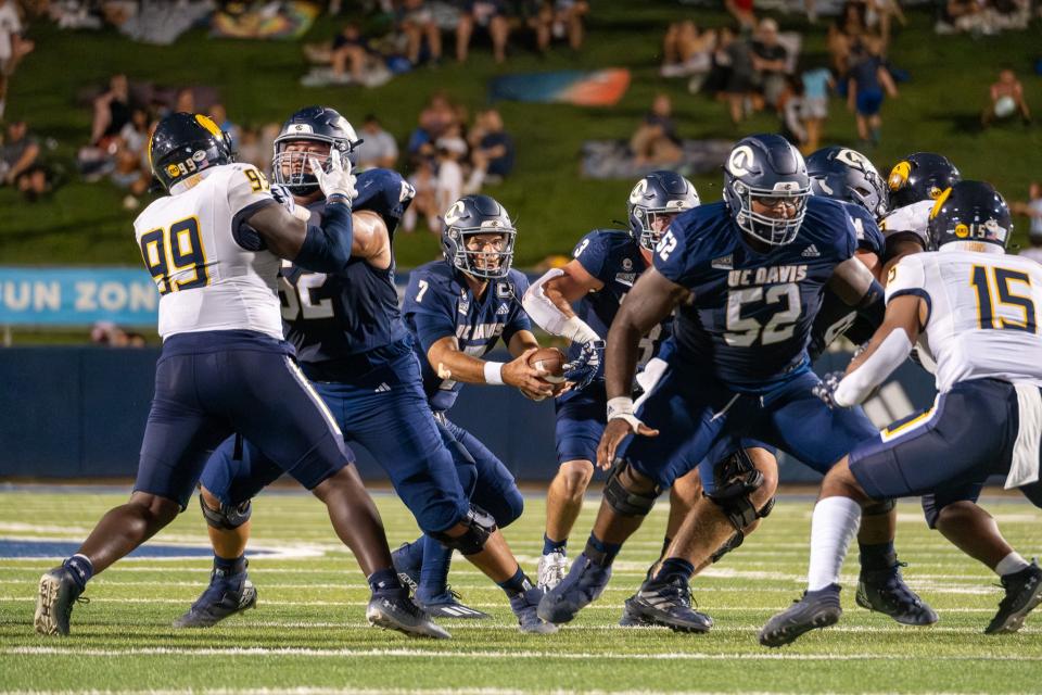 UC Davis football quarterback prepares to hand off the ball during a game against an opposing team, with players blocking and defending on the field.