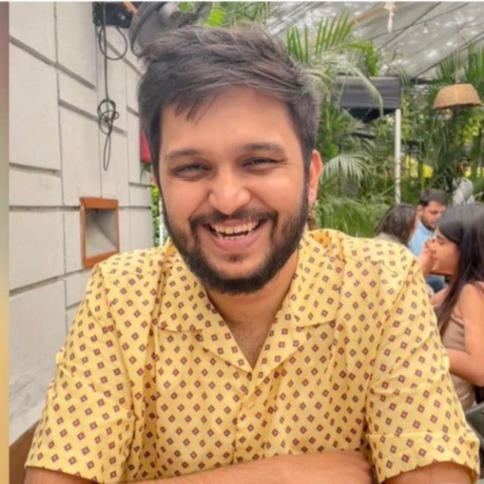 A man with short dark hair and a beard, wearing a yellow patterned shirt, sits at an outdoor cafe table, smiling at the camera. Green plants and other people are in the background.
