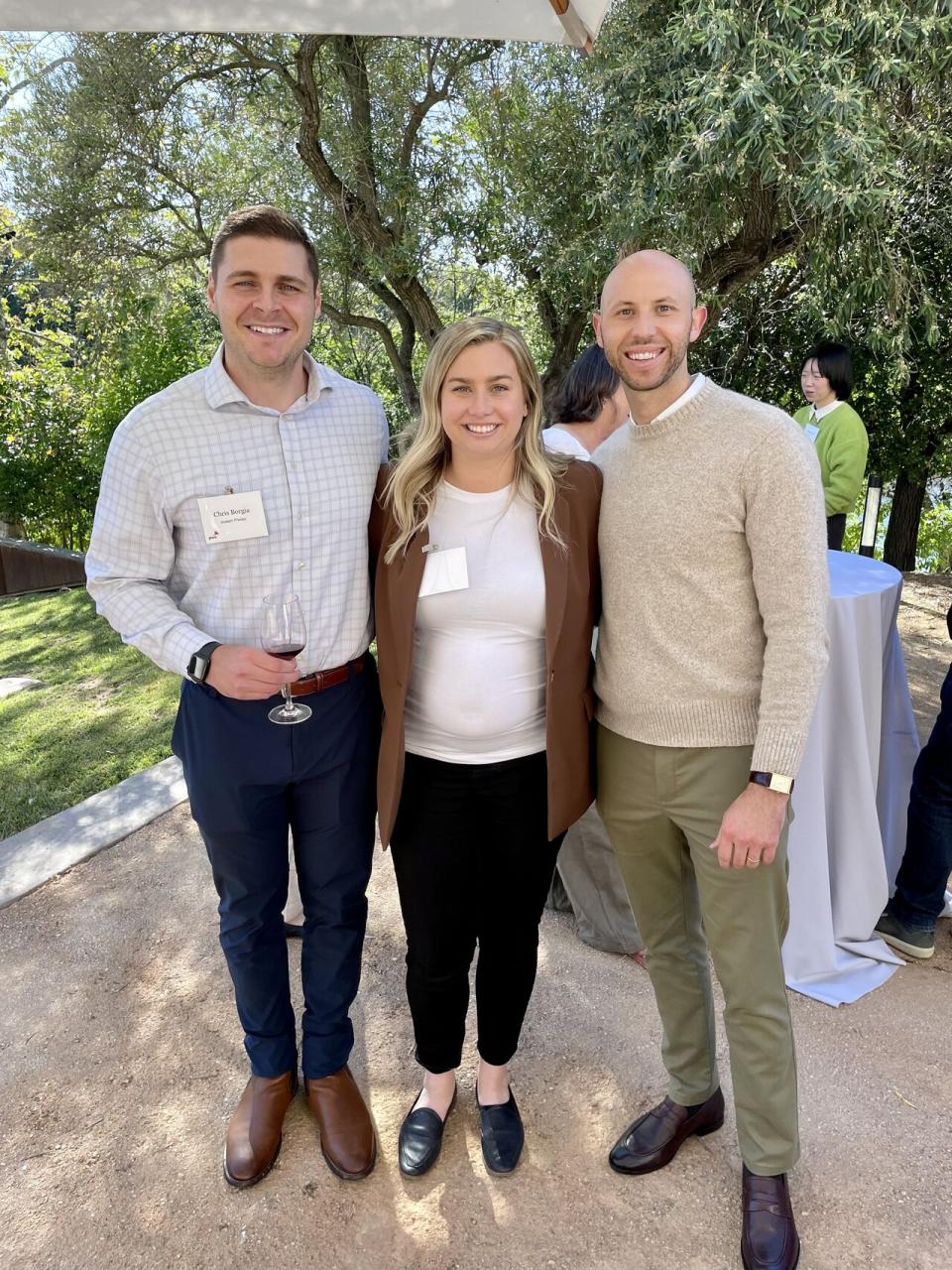 Three people stand outdoors, smiling at the camera. The two men wear business casual attire; the woman wears a white top and brown blazer. Trees and other event attendees are in the background.
