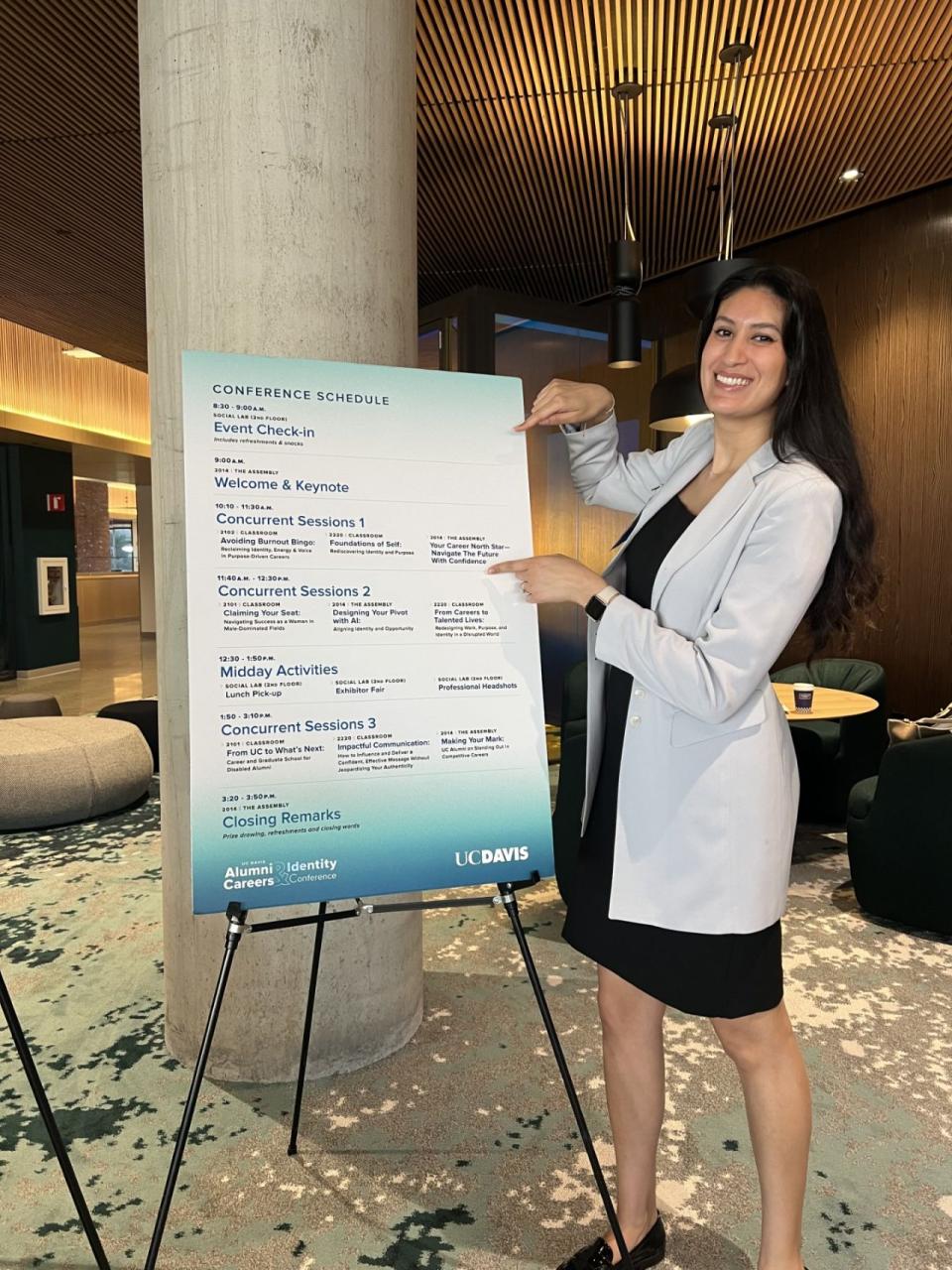 A woman in a light blazer stands indoors, smiling and pointing at a large printed conference schedule on an easel at a UC Davis event.