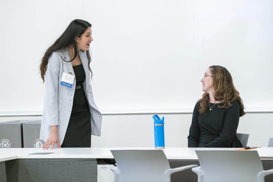 Two women interact in a modern classroom; one stands and speaks while the other sits at a desk with a blue water bottle and notebook.