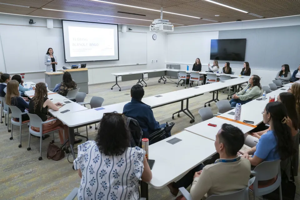A group of people sit in a classroom, facing a presenter who is giving a presentation titled "Avoiding Burnout Bingo" on a projector screen.