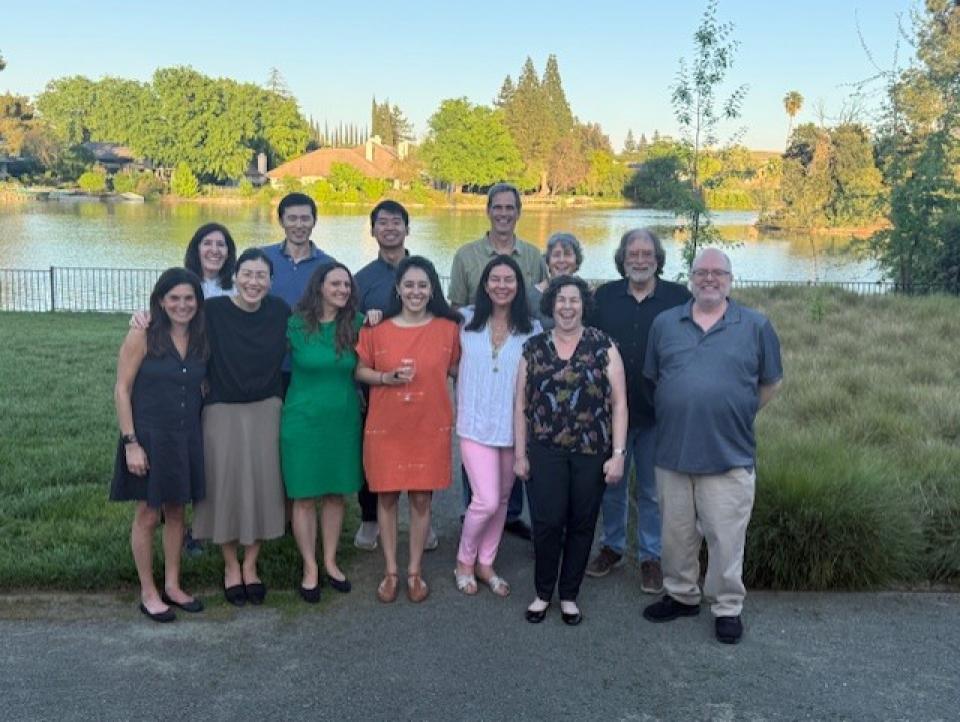 13 professors at the conference standing outdoors in front of a lake, smiling at the camera on a sunny day.