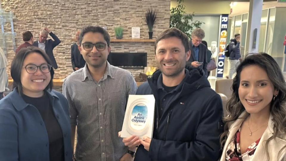 Four people stand indoors, smiling at the camera; one person in the middle holds a book titled "An Airline Odyssey." Other people are seen in the background near a reception area.