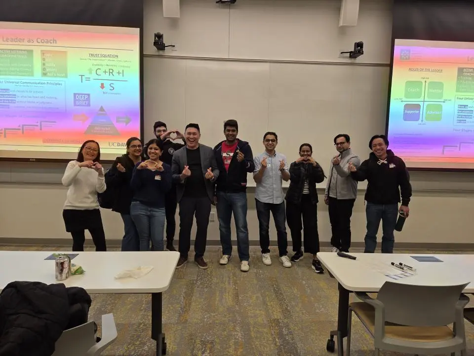 A group of ten people stand and pose together at the front of a classroom, with presentation slides displayed on screens behind them.