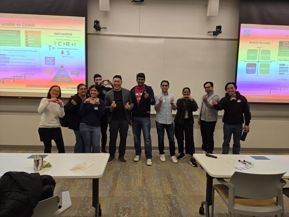 A group of ten people stand and pose together at the front of a classroom, with presentation slides displayed on screens behind them.