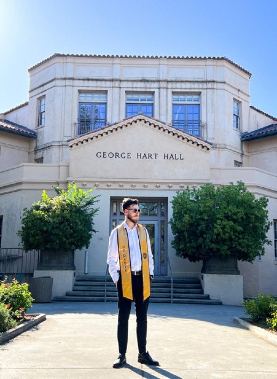 A man wearing a graduation stole stands in front of George Hart Hall on a sunny day.