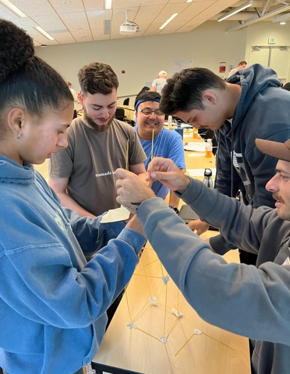 Five people work together to build a tall structure using uncooked spaghetti and marshmallows in a classroom setting.