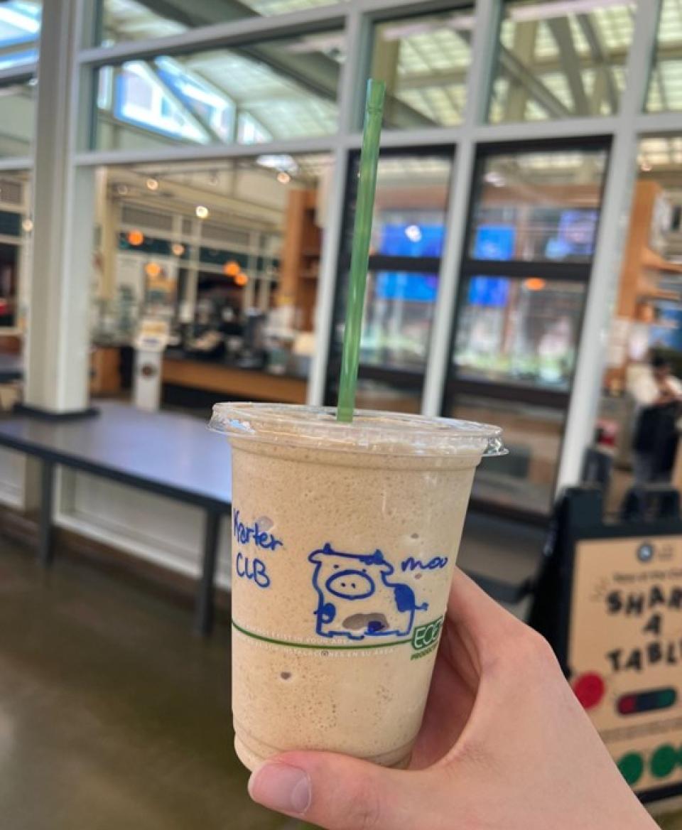 A hand holds a plastic cup filled with a blended iced drink, featuring a green straw and a drawing of a cow on the cup, inside a café with large windows.