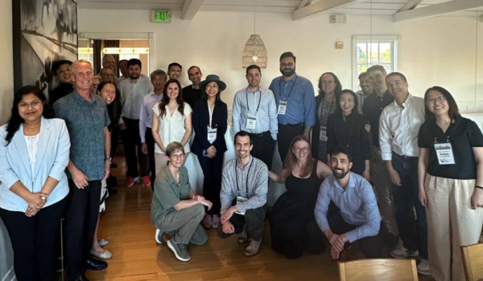 A group of about 20 people pose together indoors, some standing and some kneeling, in a well-lit room with wooden floors and white walls.