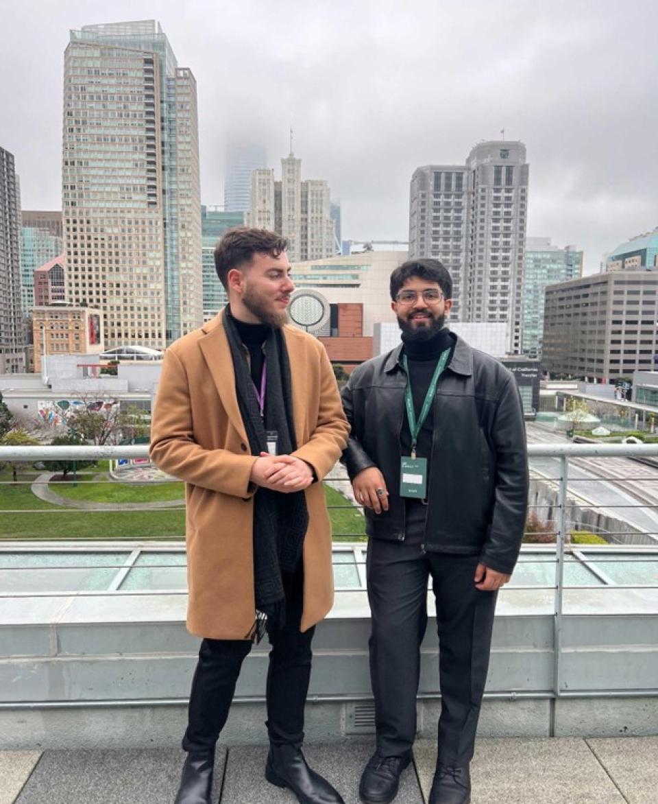 Two men stand on a rooftop terrace with city skyscrapers in the background, both wearing name badges and winter clothing.