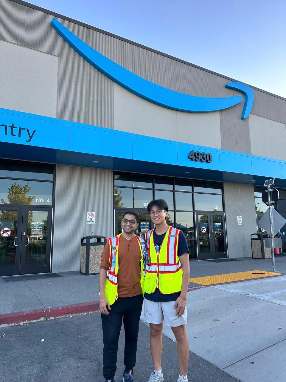 Two people wearing safety vests stand in front of an Amazon building entrance with the Amazon logo and address 4930 visible above them.