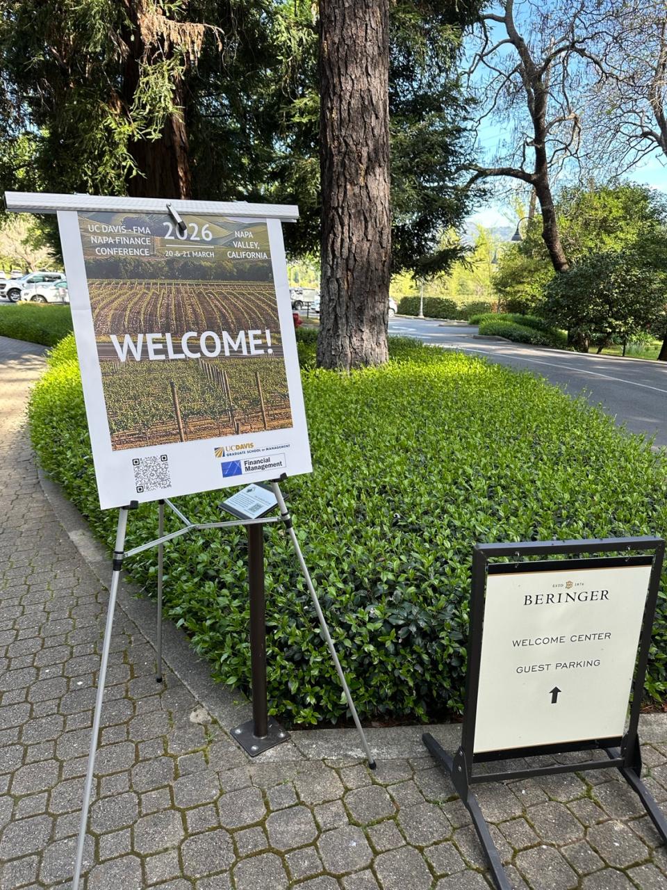 Two signs stand on a brick path; one advertises the UC Davis RNA Informatics conference, the other directs guests to Beringer Welcome Center parking, with trees and greenery in the background.
