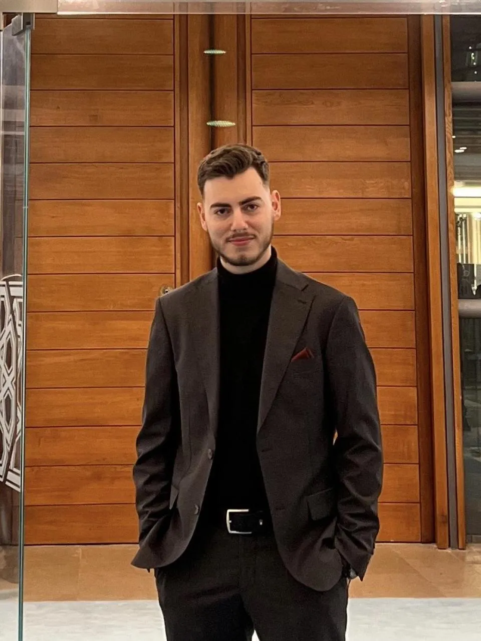 A man in a dark suit and black turtleneck stands indoors in front of wooden panel doors, with his hands in his pockets, facing the camera.