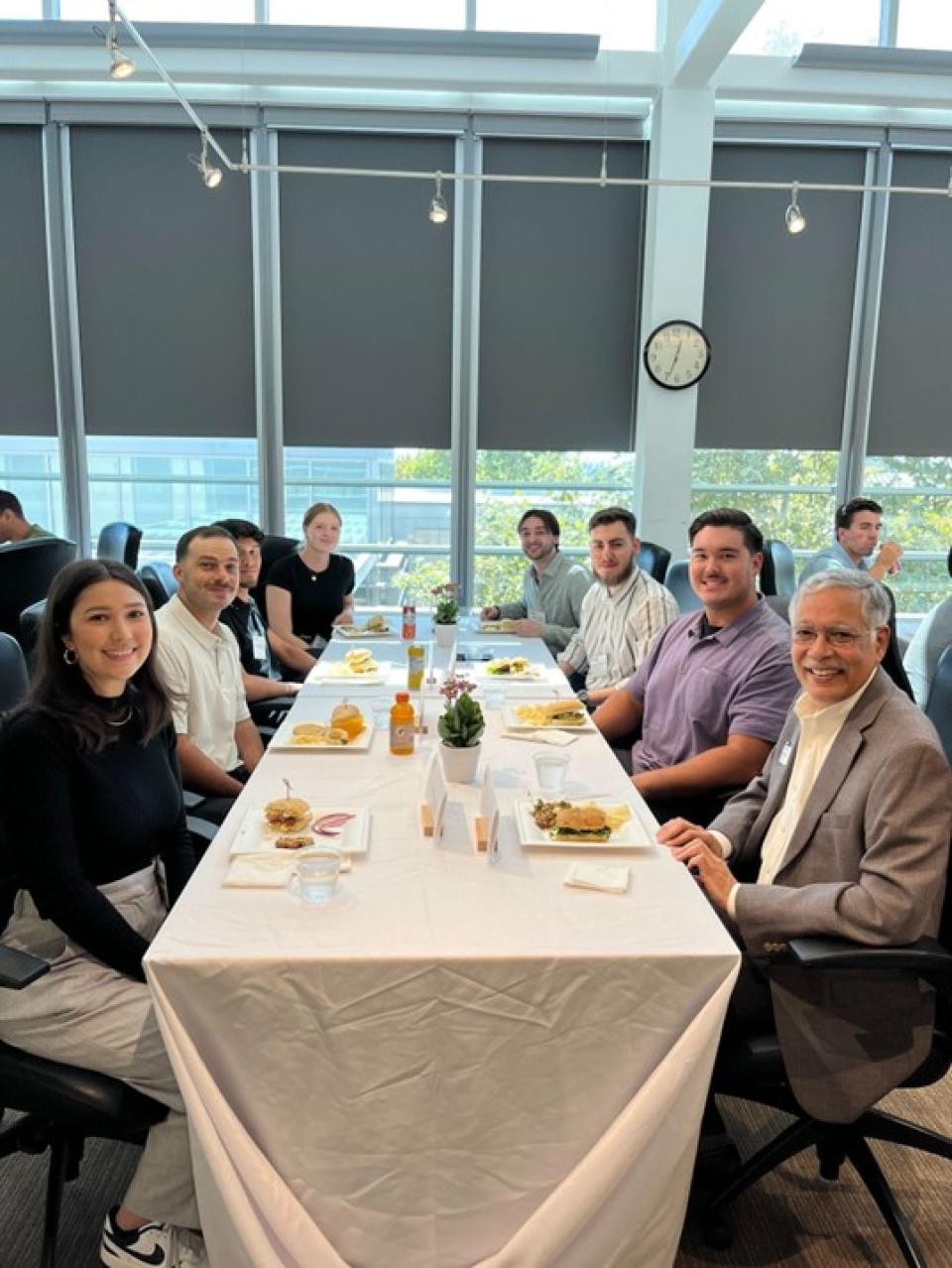 A group of people sit around two tables covered with white tablecloths, sharing a meal in a well-lit room with large windows and a clock on the wall.