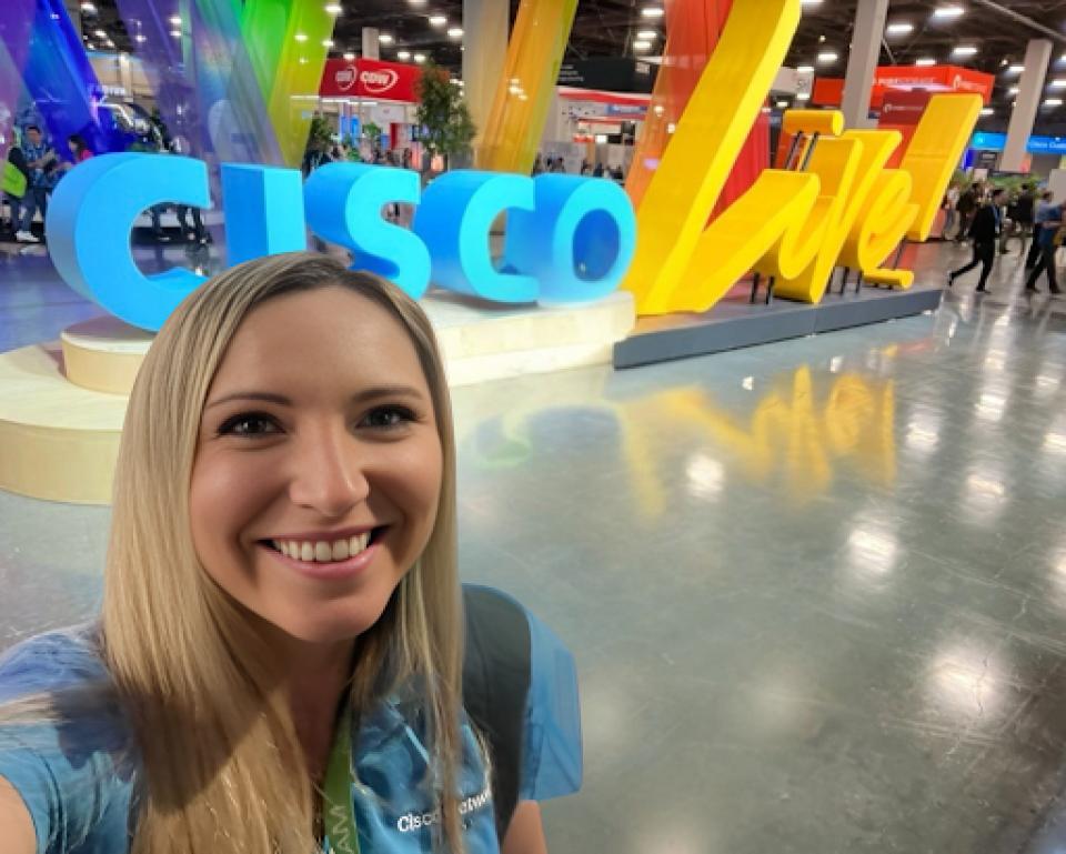 A woman takes a selfie in front of a large "CISCO Live" sign at a convention or expo, with colorful decorations and other attendees in the background.