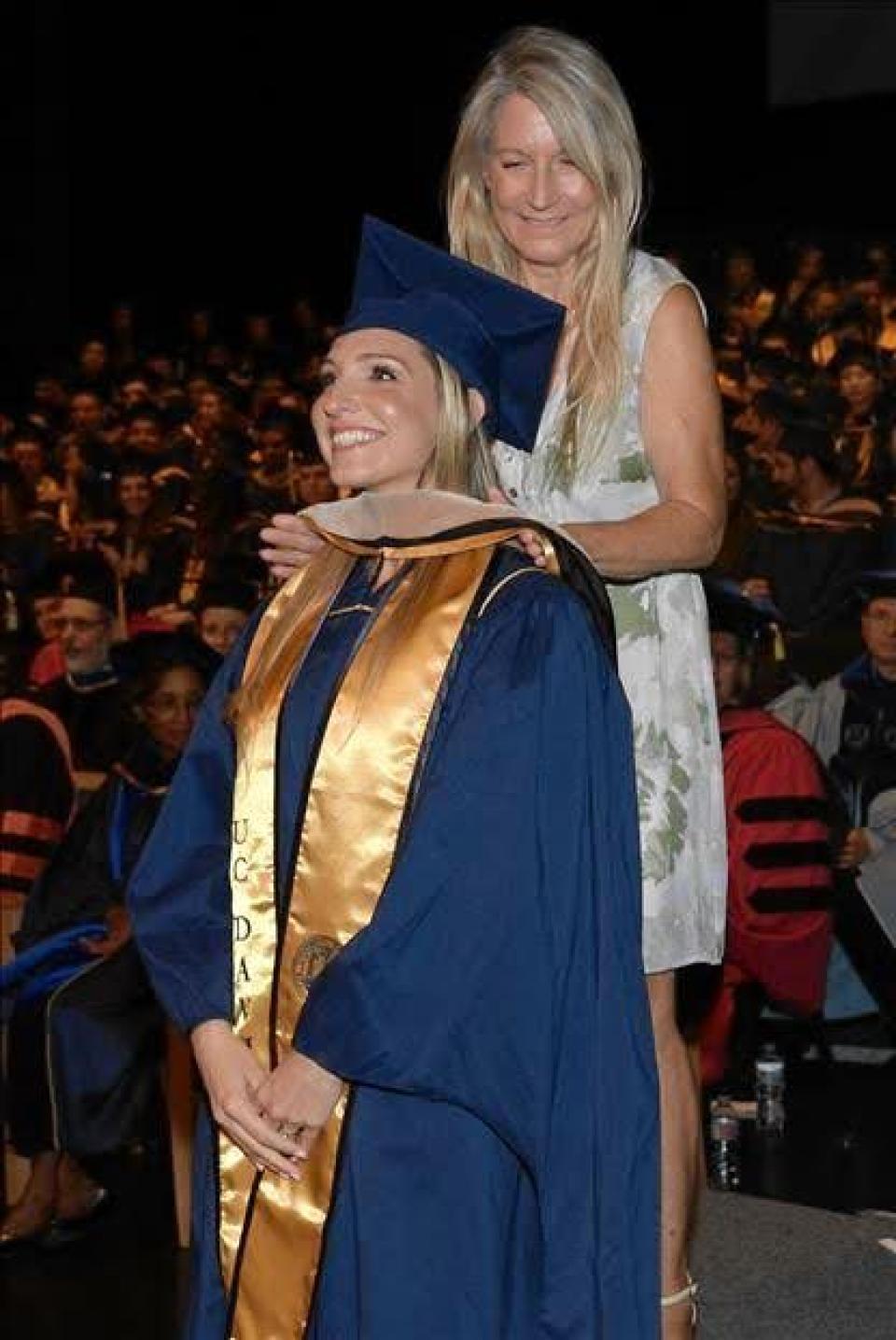 A woman in a graduation cap and gown is being hooded on stage by another woman during a commencement ceremony, with an audience in the background.