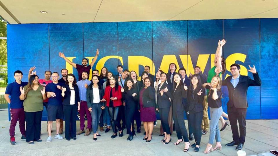 A group of people posing and smiling in front of a large blue and yellow “UC DAVIS” sign, with several making enthusiastic gestures.