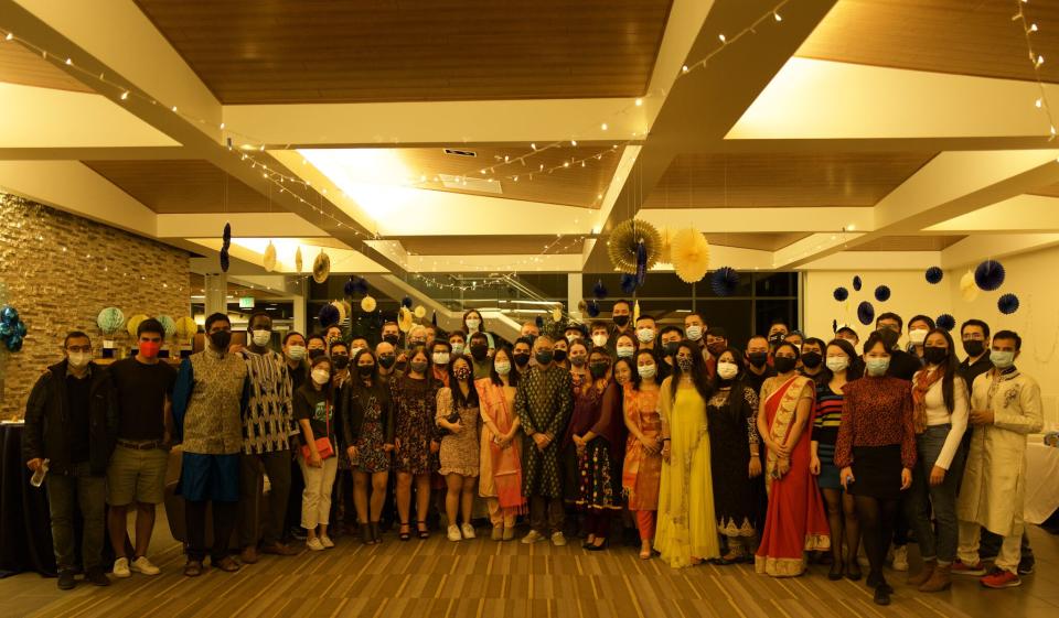 A large group of people, many in colorful traditional attire and face masks, pose together indoors under string lights and blue-yellow decorations.
