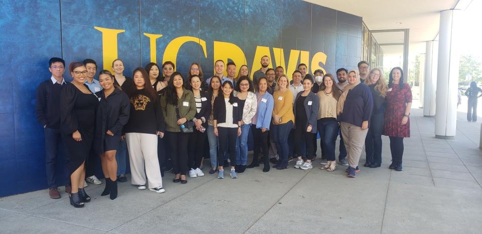 A group of people pose for a photo in front of a blue and yellow UC Davis wall on a sidewalk.