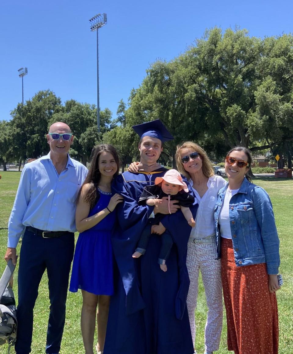 A group of five adults and a baby pose together outdoors on a grassy field. One adult is wearing a graduation cap and gown and holding the baby.