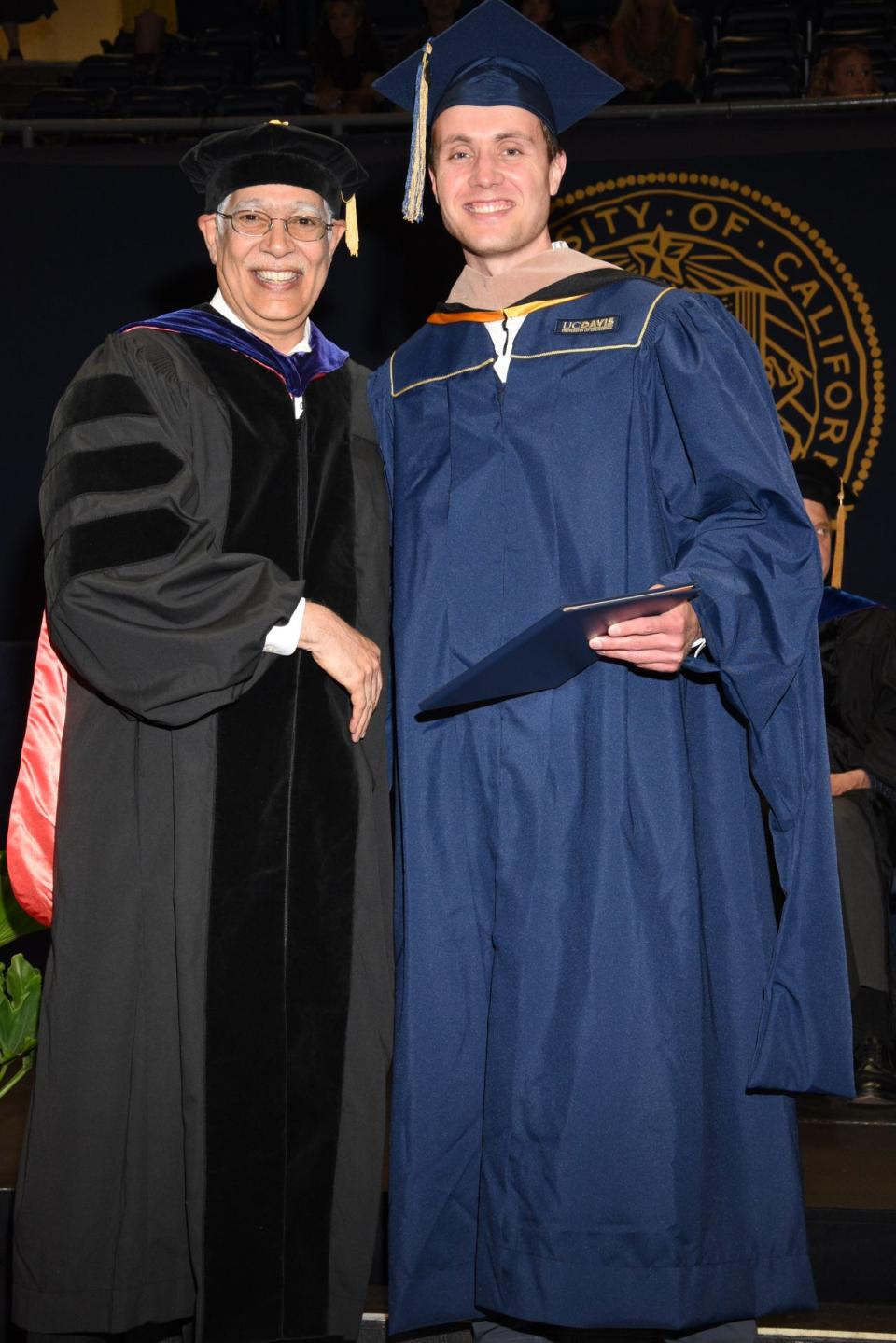 A graduate in cap and gown poses with a faculty member in academic regalia on stage at a graduation ceremony.