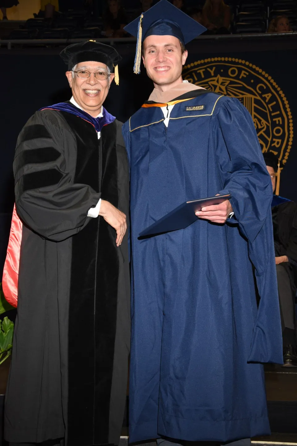 A graduate in cap and gown poses with a faculty member in academic regalia on stage at a graduation ceremony.