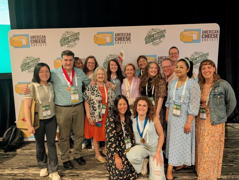 A group of people pose together in front of an American Cheese Society backdrop, many wearing name badges and some displaying medals.