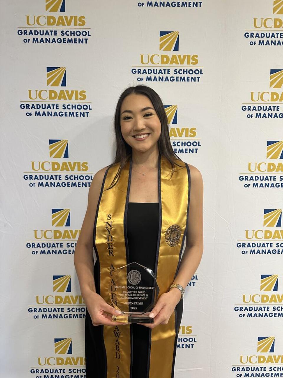 A woman in graduation attire holds an award in front of a UC Davis Graduate School of Management backdrop.