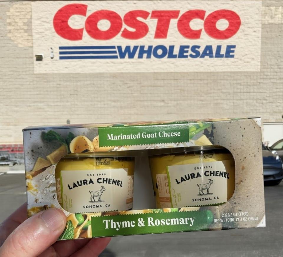 A hand holds a package of Laura Chenel Marinated Goat Cheese, Thyme & Rosemary flavor, in front of a Costco Wholesale store.
