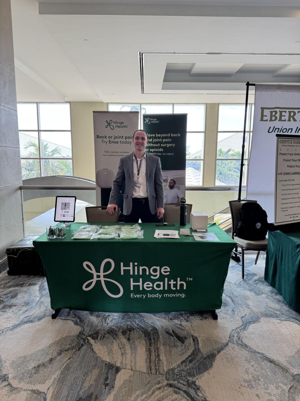 A man stands behind a Hinge Health booth with promotional materials on the table, a banner behind him, and an iPad display set up in a conference setting.