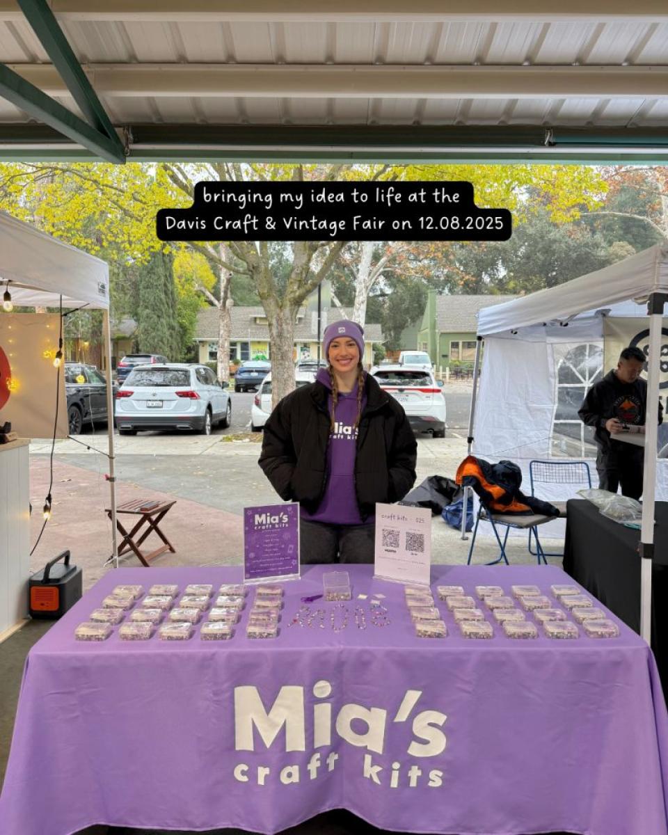 A vendor stands behind a table displaying craft kits labeled "Mia's" at an outdoor fair, with a sign about the Davis Craft & Vintage Fair on 12.08.2025.