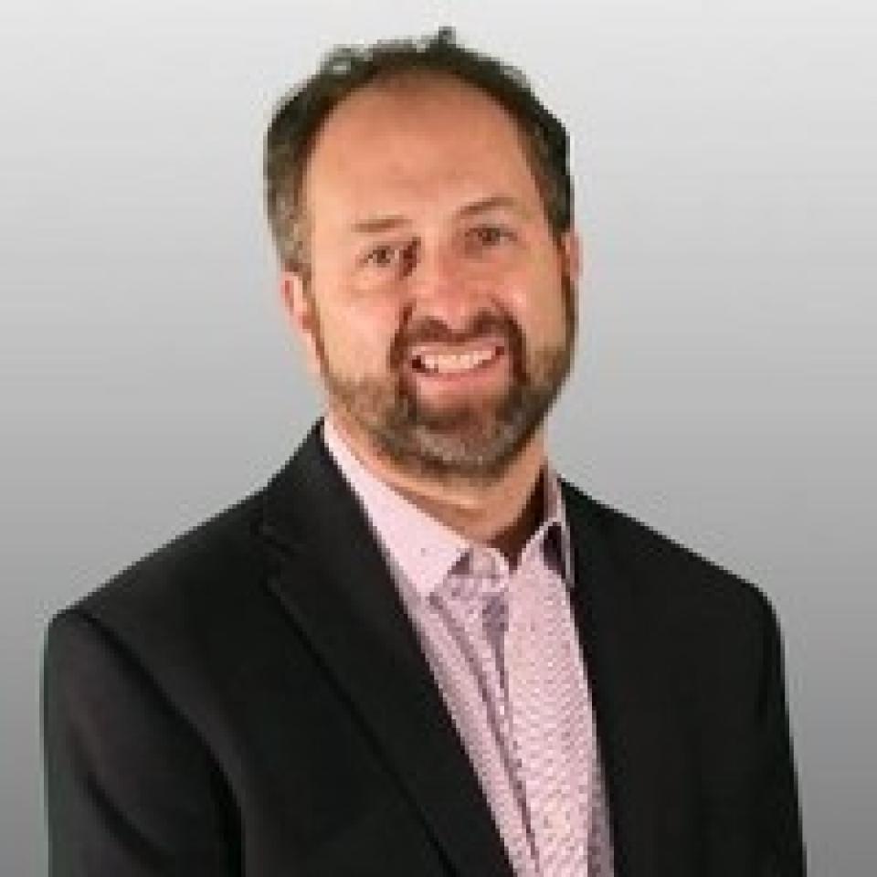 Adam Schwartz with short brown hair and a beard, wearing a dark suit jacket over a light pink patterned shirt, stands in front of a plain gray background, smiling at the camera.