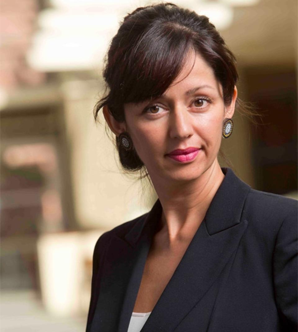 Amelia Miazad , wearing a black blazer and earrings, stands outdoors with a blurred building background.