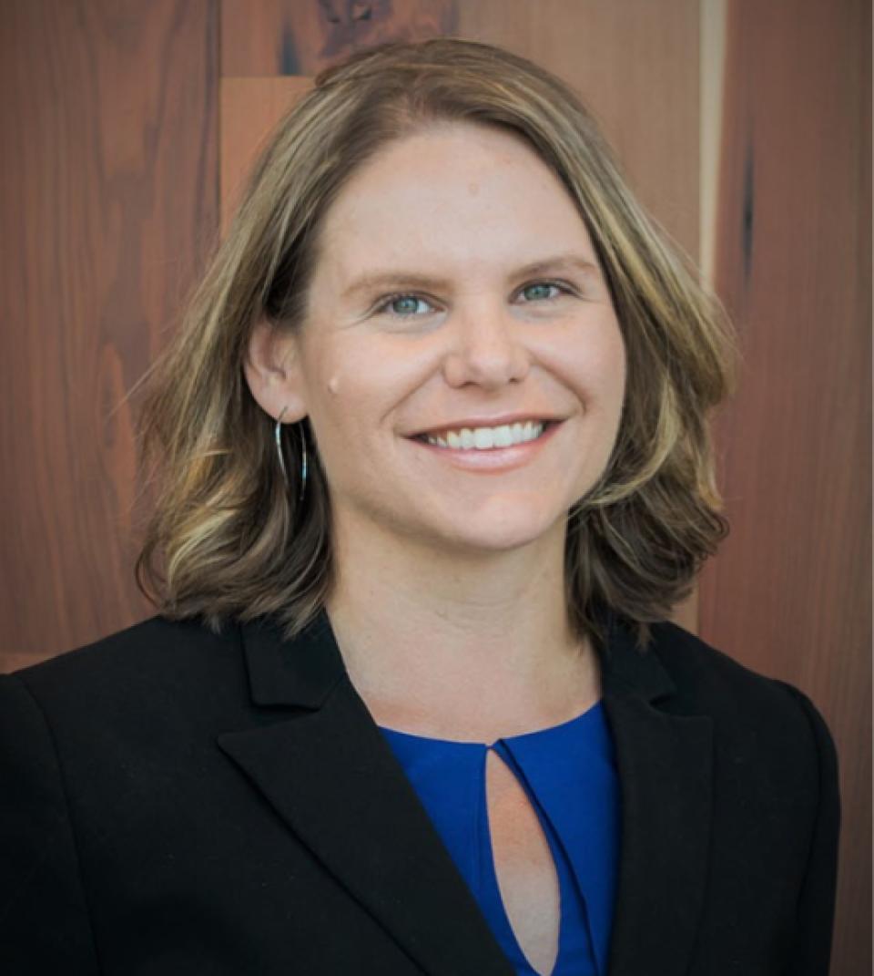 Anne Visser, wearing a black blazer over a blue blouse, smiles in front of a wooden background.