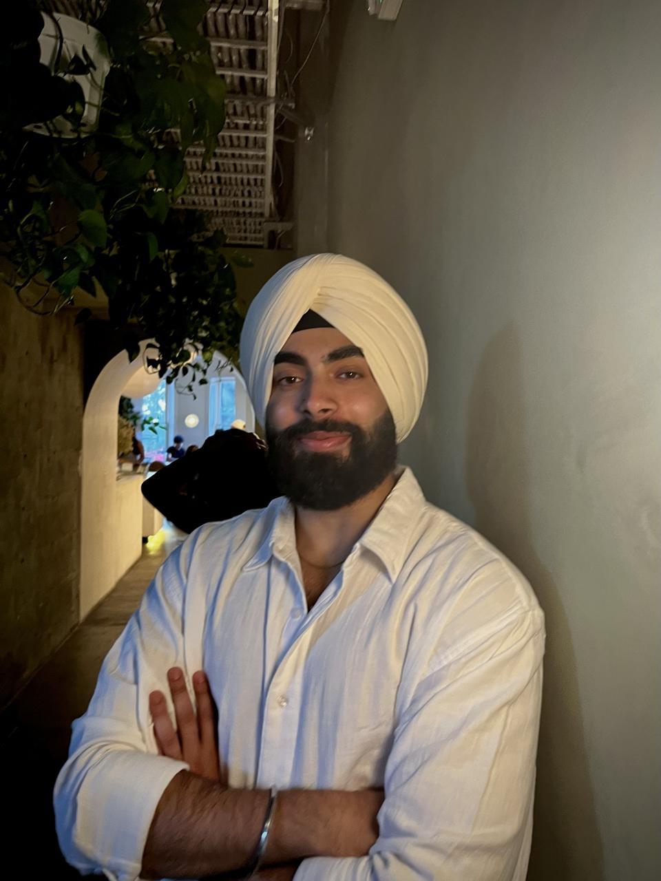 Gunraj Singh stands with arms crossed, smiling slightly, in a dimly lit hallway with plants and arched doorways in the background.