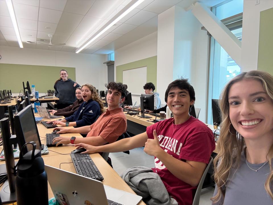 A group of students sit at desks with computers in a classroom, smiling at the camera; an instructor stands at the back of the room.