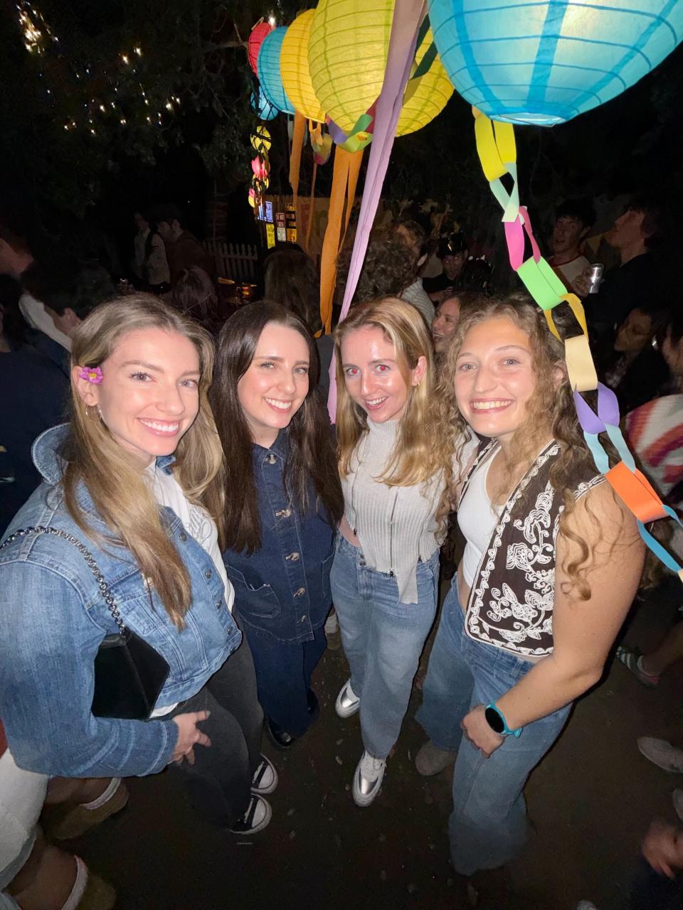 Four young women stand close together smiling at an outdoor nighttime party, surrounded by colorful paper lanterns and hanging decorations.