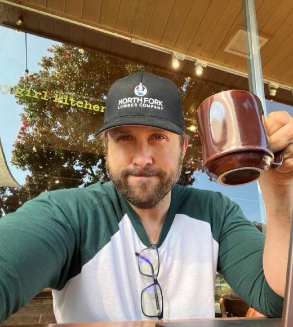 Keith Taylor wearing a baseball cap and a green and white shirt holds up a large brown coffee mug while sitting at an outdoor café table.