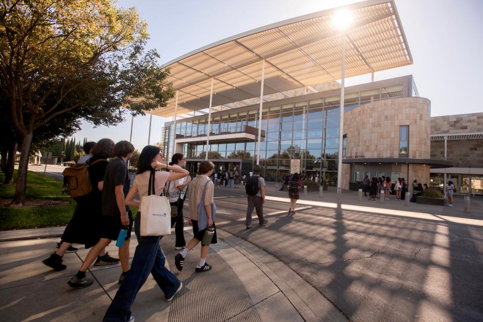 A group of people walk toward a modern glass building with a large overhanging roof on a sunny day.