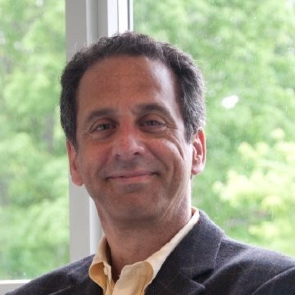 Richard Morris in a dark blazer and light shirt smiles while seated indoors in front of a window with green trees visible outside.
