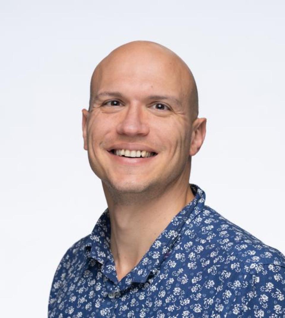 Seth Frey smiles at the camera, wearing a blue shirt with a white floral pattern, against a plain light background.
