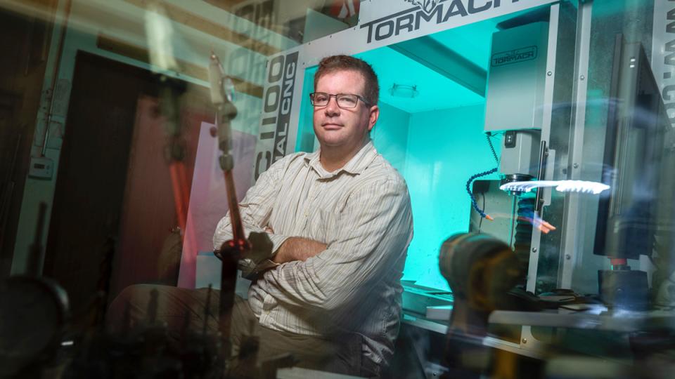 A man wearing glasses and a striped shirt sits with arms crossed in front of a Tormach CNC machine in a workshop.