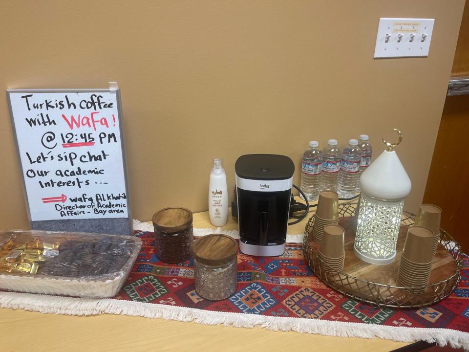 A table with Turkish coffee setup, snacks, a coffee maker, water bottles, a lantern, and a sign announcing an academic coffee chat event with Wafa at 12:45 PM.