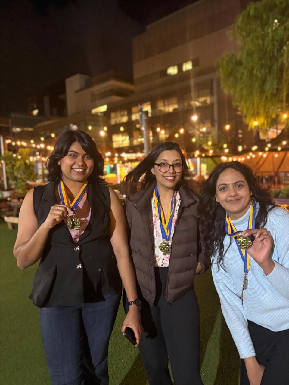 Three women stand outdoors at night, smiling and holding up medals on ribbons around their necks, with string lights and buildings in the background.