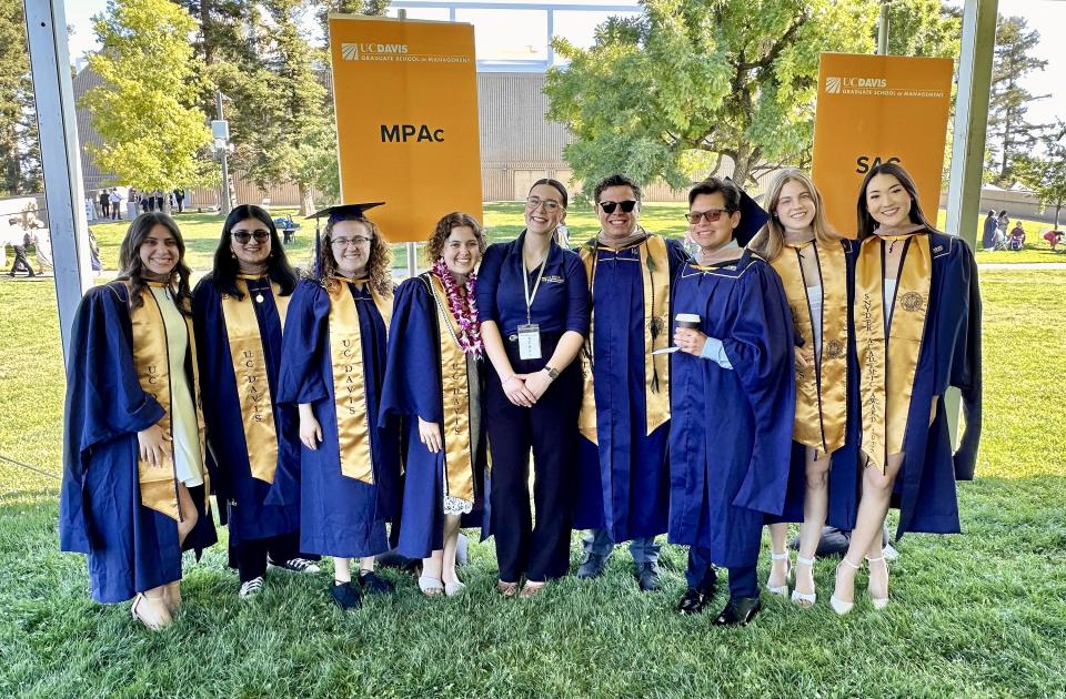 A group of graduates in caps and gowns stand together outdoors in front of UC Davis banners for MPAc and SAC, smiling at the camera.