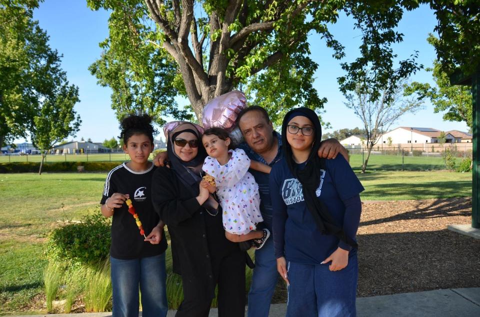 A family of five poses together outdoors in a park on a sunny day, with trees, grass, and houses in the background. One member holds heart-shaped balloons.