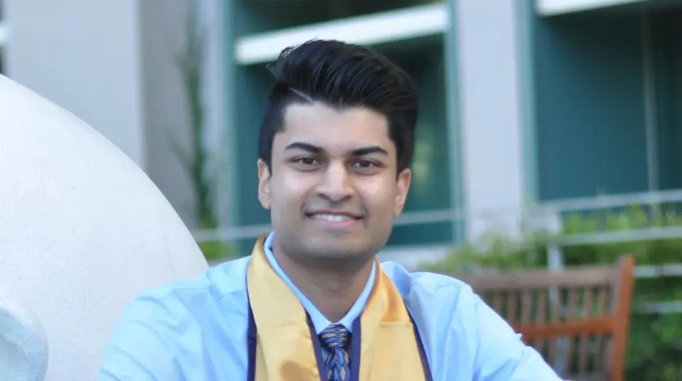 A person wearing a light blue shirt, patterned tie, and a gold graduation stole is smiling outdoors in front of a building.